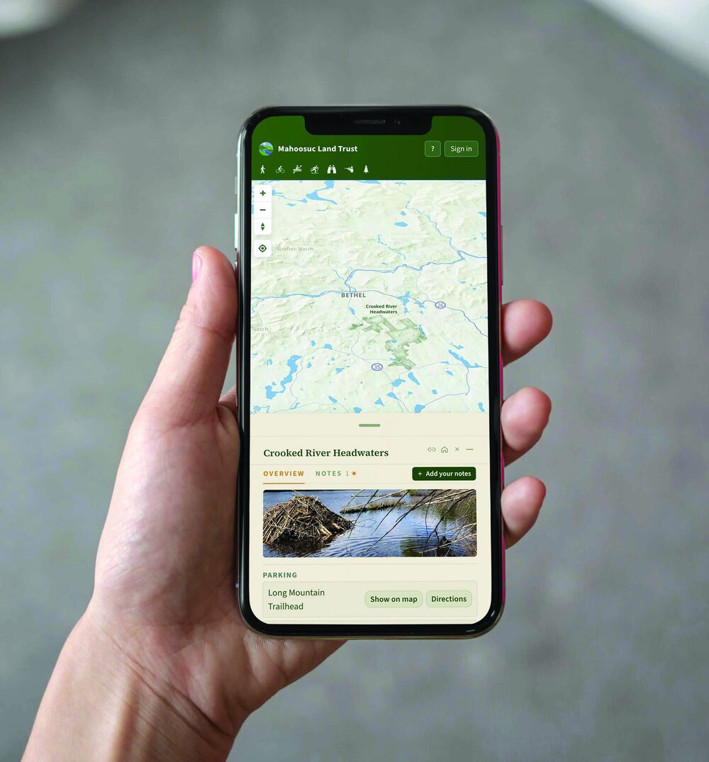 A hand holding a phone showing the Mahoosuc Land Trust map, with the Crooked River Headwaters preserve selected